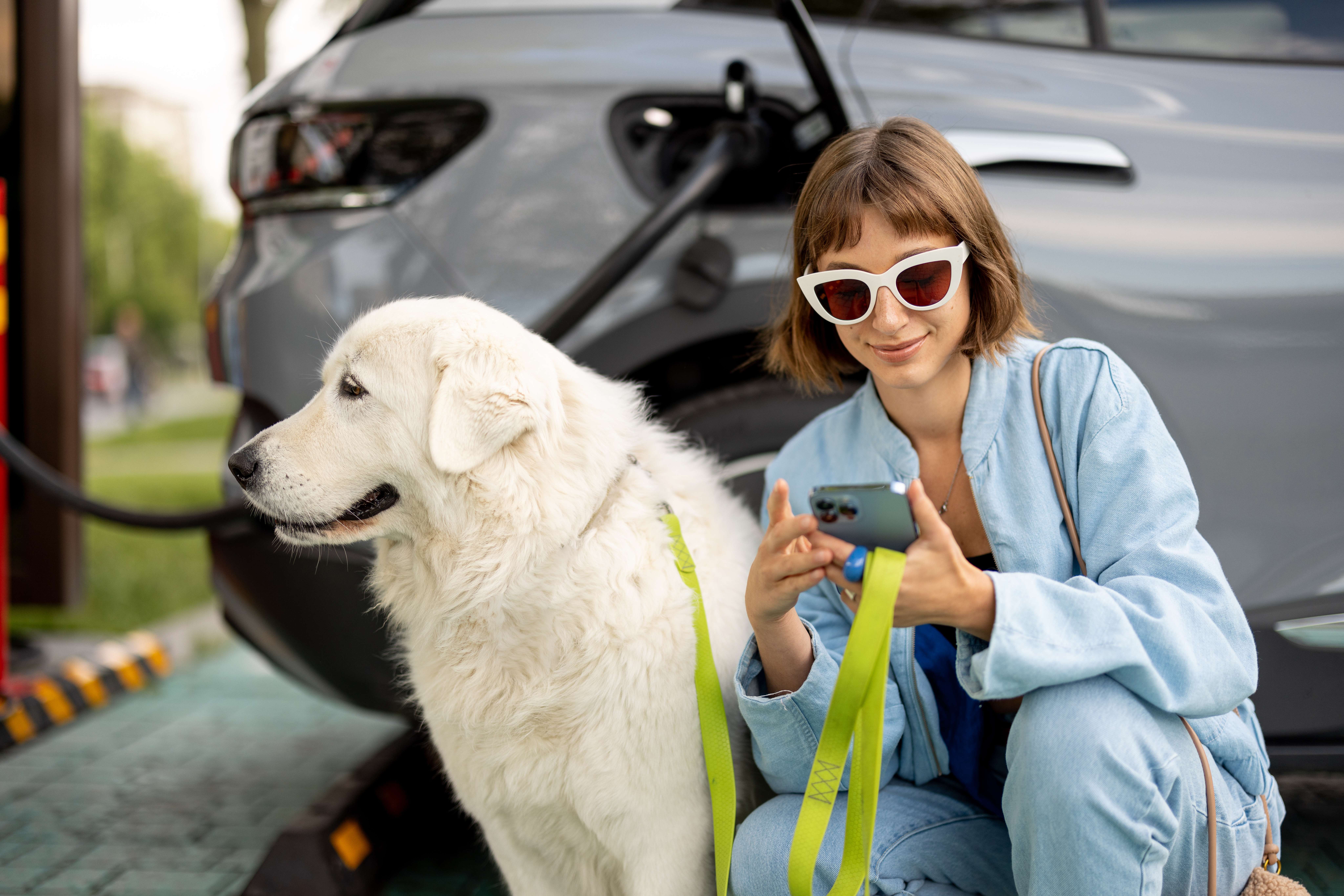 Woman With Dog Waiting For Electric Car To Be Charged On A Public Station